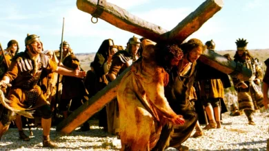 Cena impactante do filme A Paixão de Cristo (2004), dirigido por Mel Gibson, com destaque para a direção de arte e figurino autêntico. Jesus, interpretado por Jim Caviezel, caminha sob o peso da cruz em uma paisagem árida, cercado por figurantes em trajes da época.