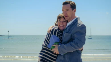 Cena do filme "Os Roses: Até Que a Morte Os Separe" (2025), com Benedict Cumberbatch e Olivia Colman abraçados na praia em clima de romance e tensão.