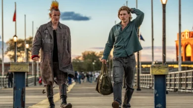 Cena do filme Ladrões (2025), com Matt Smith de moicano e casaco punk ao lado de Austin Butler, que carrega uma mochila em uma passarela ao entardecer.