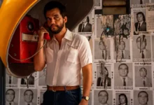 Wagner Moura em cena do filme O Agente Secreto (2025), dirigido por Kleber Mendonça Filho; o ator aparece em um orelhão vermelho, usando camisa branca, em frente a um muro coberto por cartazes políticos dos anos 1970.