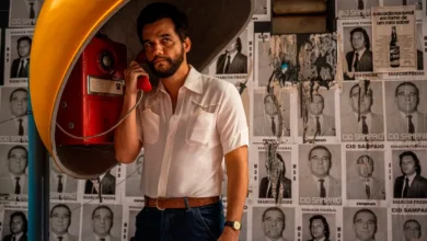 Wagner Moura em cena do filme O Agente Secreto (2025), dirigido por Kleber Mendonça Filho; o ator aparece em um orelhão vermelho, usando camisa branca, em frente a um muro coberto por cartazes políticos dos anos 1970.