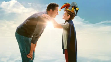 Zachary Levi e Jacob Laval em cena promocional do filme Invencível (2025); pai e filho trocam um olhar carinhoso sob um céu azul, com o menino vestindo capa e chapéu de bobo da corte.