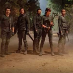 Cena de The Old Guard 2 (2025), filme de ação da Netflix, mostra grupo de guerreiros imortais liderados por Andy (Charlize Theron) em floresta sombria, todos vestidos com trajes táticos e armados, prontos para o combate.