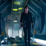 Charlize Theron como Andy em cena de ação intensa de The Old Guard 2 (2025), segurando seu machado em corredor tecnológico, após confronto violento.