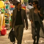 Cena de The Old Guard 2 (2025) mostra Andy (Charlize Theron) e Copley (Chiwetel Ejiofor) caminhando juntos por mercado asiático, em nova missão da equipe imortal.
