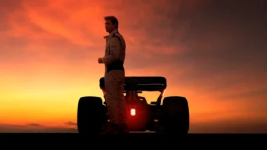 Brad Pitt em traje de piloto de corrida, silhuetado contra um pôr do sol alaranjado e dramático, de pé na frente de um carro de Fórmula 1, com as luzes traseiras vermelhas acesas.