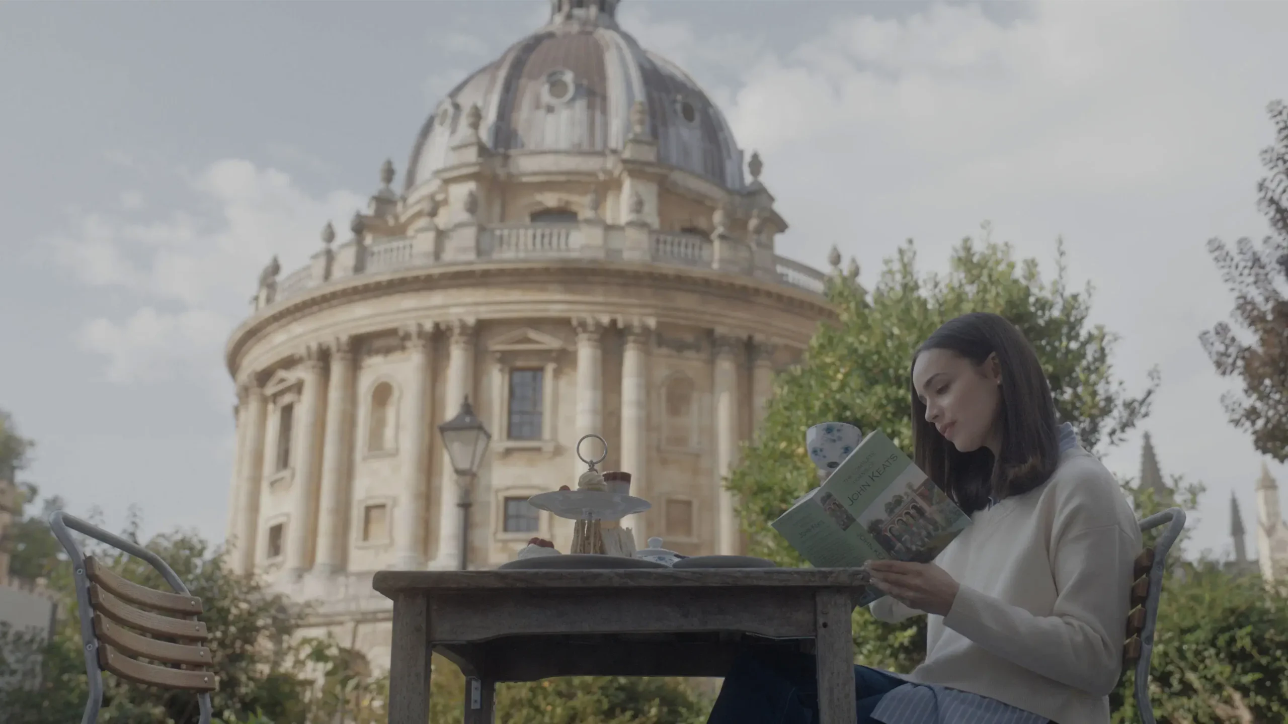 Sofia Carson lendo um livro de John Keats em uma mesa ao ar livre, com a Radcliffe Camera ao fundo, em cena do filme Meu Ano em Oxford (2025)