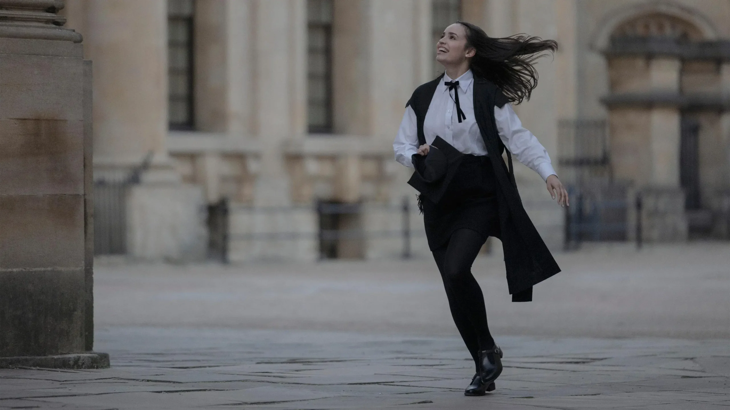 Sofia Carson corre sorridente pelas ruas de Oxford, vestindo traje acadêmico tradicional, em cena do filme Meu Ano em Oxford (2025)