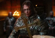 Cena do filme Drácula: Uma História de Amor Eterno, com Caleb Landry Jones usando armadura medieval e segurando uma cruz dourada.