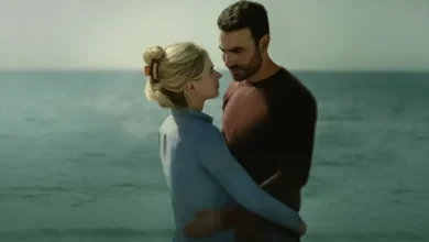 Uma cena do filme de romance 'Por Inteiro' (All of You) com Imogen Poots e Brett Goldstein. Imogen Poots (loira) e Brett Goldstein (moreno) se abraçam e se olham intensamente à beira da água, com o oceano ao fundo.