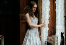 Atriz Lea Myren como Elvira em cena de A Meia-Irmã Feia (2025), filme de terror dirigido por Emilie Blichfeldt, observando pela janela em uma casa antiga com cortinas rendadas.