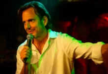 Bradley Cooper segurando microfone e sorrindo em cena de Isso Ainda Está de Pé dirigida por ele mesmo