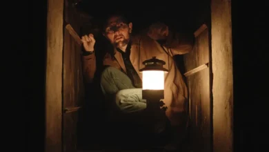 Adam Scott em cena sombria de Hokum: O Pesadelo da Bruxa (2026), segurando uma lanterna dentro de um espaço escuro e claustrofóbico.
