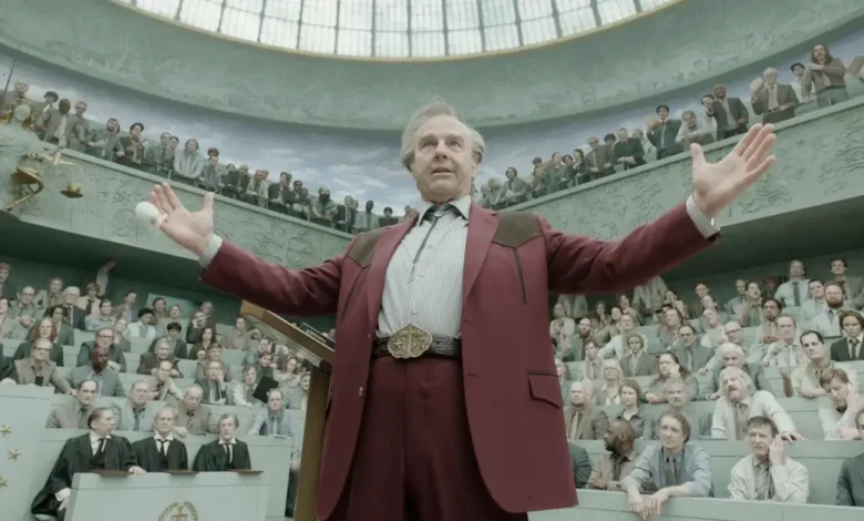 Tom Cruise em cena do Trailer de Digger, filme dirigido por Alejandro G. Iñárritu, durante um discurso em um grande tribunal cercado por uma multidão de espectadores.