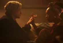 Tom Rhys Harries e Naomi Ackie fazem parte do elenco de Cara-de-Barro (2026) em cena dramática do filme, com os personagens frente a frente em um quarto sob iluminação quente.