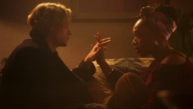 Tom Rhys Harries e Naomi Ackie fazem parte do elenco de Cara-de-Barro (2026) em cena dramática do filme, com os personagens frente a frente em um quarto sob iluminação quente.