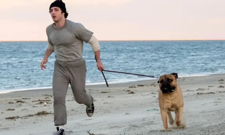 Anthony Ippolito como Sylvester Stallone corre pela praia durante treinamento em trailer de Eu Sou o Rocky (I Play Rocky, 2026), acompanhado de um cachorro.