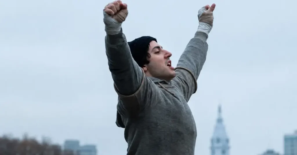 Anthony Ippolito como Sylvester Stallone celebra com os braços erguidos em cena de Eu Sou o Rocky (I Play Rocky, 2026) inspirada na icônica sequência de treinamento de Rocky.