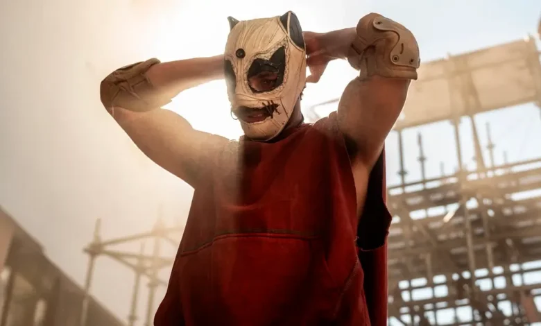 Personagem mascarado em cena de Corrida dos Bichos (2026), thriller de ação dirigido por Fernando Meirelles, em ambiente industrial tomado por fumaça.