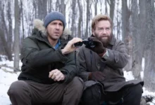 Kenneth Branagh e Ryan Reynolds observam a distância com binóculos em uma floresta coberta de neve em uma cena de Mayday (2026).
