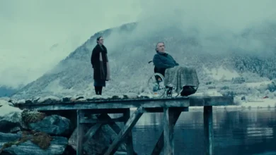 Erik Hivju e Renate Reinsve em uma cena do filme Fjord (2026), ao lado de um fiorde cercado por montanhas cobertas de neve.