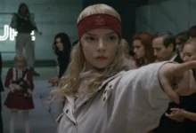 Anya Taylor-Joy em cena do filme Sacrifício (2026), usando uma faixa vermelha na cabeça e apontando para alguém durante um evento diante de espectadores.
