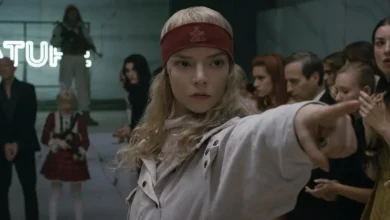 Anya Taylor-Joy em cena do filme Sacrifício (2026), usando uma faixa vermelha na cabeça e apontando para alguém durante um evento diante de espectadores.