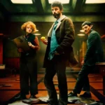 Imagem promocional da série Departamento Q da Netflix, com os atores Leah Byrne, Matthew Goode (ao centro, segurando uma bola de tênis) e Alexej Manvelov em um ambiente sombrio de escritório com iluminação dramática. Estreia em 29 de maio de 2025.