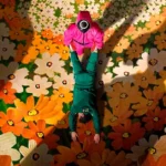 Cena promocional da terceira temporada de Round 6 com guarda mascarado em rosa segurando participante com uniforme verde, sobre fundo floral.