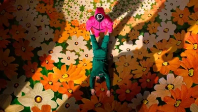 Cena promocional da terceira temporada de Round 6 com guarda mascarado em rosa segurando participante com uniforme verde, sobre fundo floral.