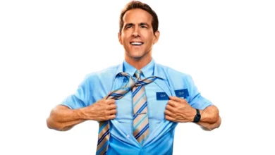 Ryan Reynolds como Guy em Free Guy, sorrindo enquanto abre sua camisa azul de trabalho para revelar uma camiseta azul com a palavra "GUY" escrita, usando gravata listrada em tons de azul e laranja