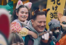 "Brendan Fraser carregando criança nos ombros em cena do filme Família de Aluguel, cercados por pessoas vestidas com fantasias de animais em festival japonês