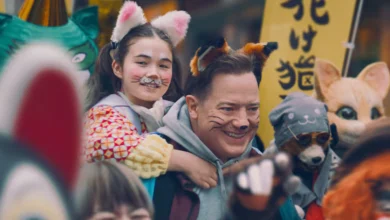 "Brendan Fraser carregando criança nos ombros em cena do filme Família de Aluguel, cercados por pessoas vestidas com fantasias de animais em festival japonês