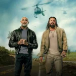 Jason Momoa e Dave Bautista em cena promocional do filme Dupla Perigosa (2026), com helicópteros ao fundo em ambiente tropical.