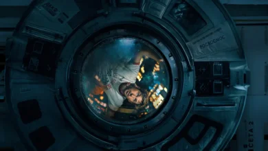 Cena do filme Devoradores de Estrelas (Project Hail Mary): astronauta visto através de escotilha circular de nave espacial, com interior iluminado ao fundo