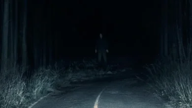 Cena sombria do filme Passageiro do Mal (2026), dirigido por André Øvredal, mostrando uma estrada escura cercada por árvores e uma figura misteriosa ao fundo.