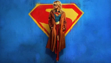 Milly Alcock como Supergirl no filme Supergirl (2026), usando um sobretudo marrom sobre o uniforme da heroína, diante do icônico símbolo da Casa de El em um fundo azul.