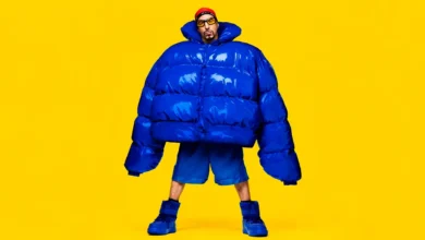 Sacha Baron Cohen caracterizado como Ali G no filme Ali G: Who Iz I? (2026), usando jaqueta azul oversized, óculos amarelos e gorro vermelho sobre fundo amarelo.