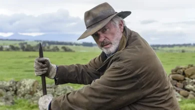 Cena do Trailer de Os Fazendeiros (Rams, 2020) mostra Sam Neill em uma fazenda rural, usando chapéu e segurando uma ferramenta de trabalho durante cena do drama.