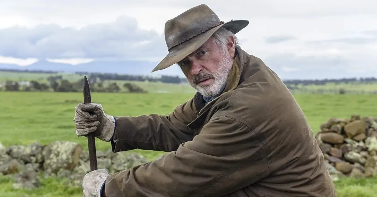 Cena do Trailer de Os Fazendeiros (Rams, 2020) mostra Sam Neill em uma fazenda rural, usando chapéu e segurando uma ferramenta de trabalho durante cena do drama.