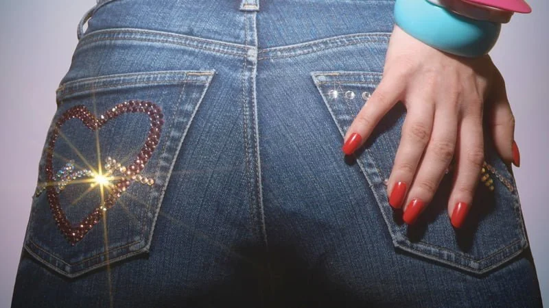 Parte traseira de uma calça jeans decorada com strass em forma de coração, mão com unhas vermelhas e pulseiras coloridas, imagem promocional do filme de terror cômico Slaxx (2020).