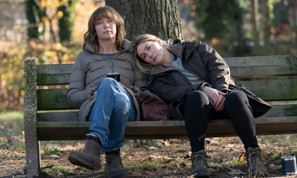Kate Winslet e Angourie Rice sentadas em banco de parque em cena de Mare of Easttown da HBO