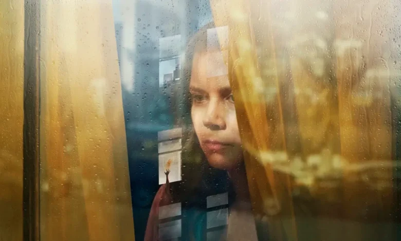 Amy Adams em cena do thriller psicológico A Mulher na Janela (2021), observando pela janela em meio a reflexos e clima de suspense.