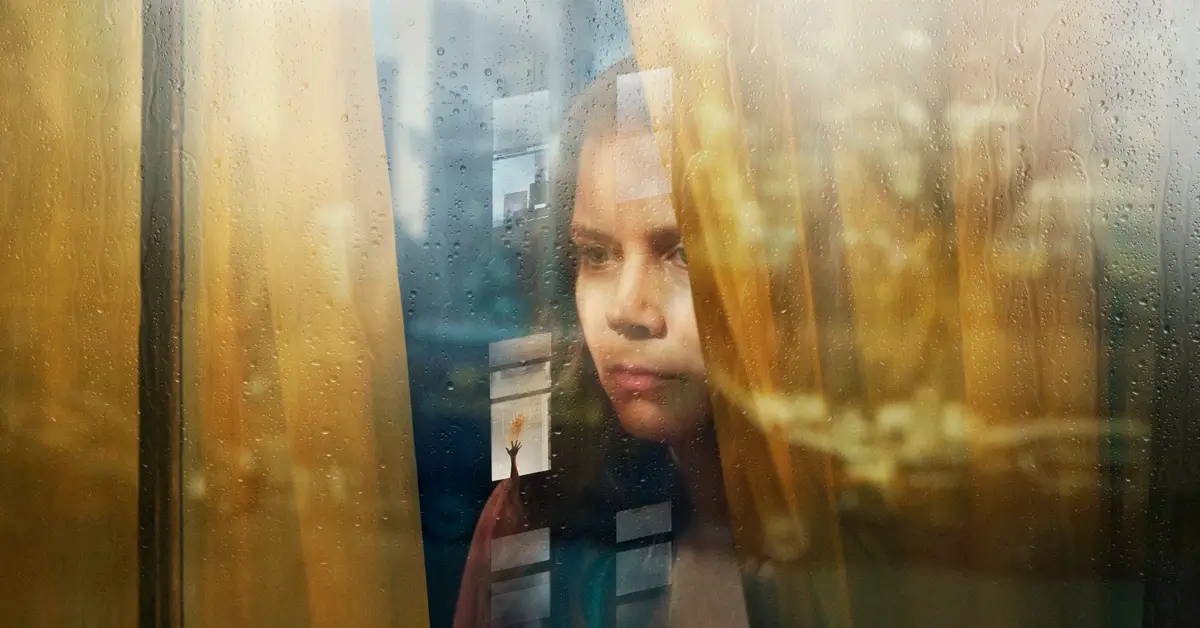 Amy Adams em cena do thriller psicológico A Mulher na Janela (2021), observando pela janela em meio a reflexos e clima de suspense.