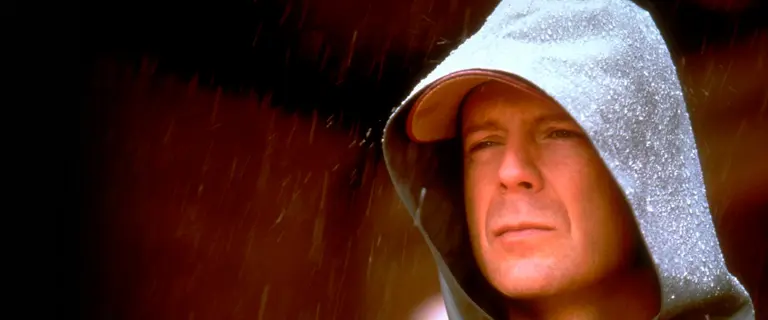 Bruce Willis em cena do filme Corpo Fechado de 2000, mostrando seu personagem com expressão séria e usando um boné em uma cena marcante do filme.