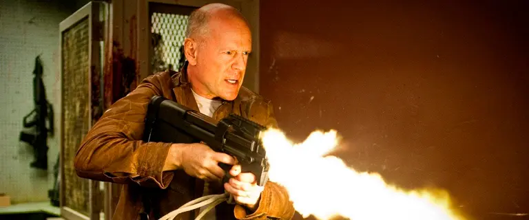 Bruce Willis no filme Looper: Assassinos do Futuro, segurando uma arma e atirando, com expressão intensa e cenário futurista ao fundo.