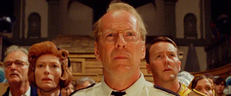 Bruce Willis no filme Moonrise Kingdom (2012), atuando com expressão séria em uma cena de filme com outros personagens ao fundo.