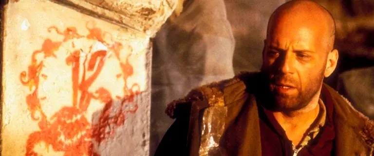 Bruce Willis interpretando um personagem em Os 12 Macacos (1995), com expressão séria ao lado de uma parede com um relógio antigo. Cena do filme de ficção científica dirigido por Terry Gilliam.