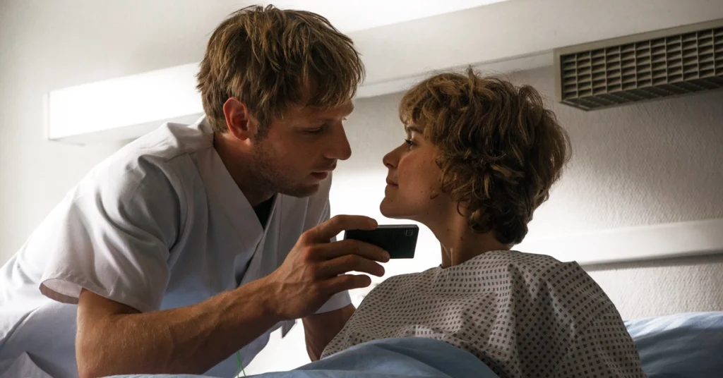 Cena do filme Wetlands (2013), com os atores Christoph Letkowski e Carla Juri; ele veste uniforme de enfermagem e segura um celular próximo ao rosto dela, que está deitada em uma cama de hospital com expressão intensa.