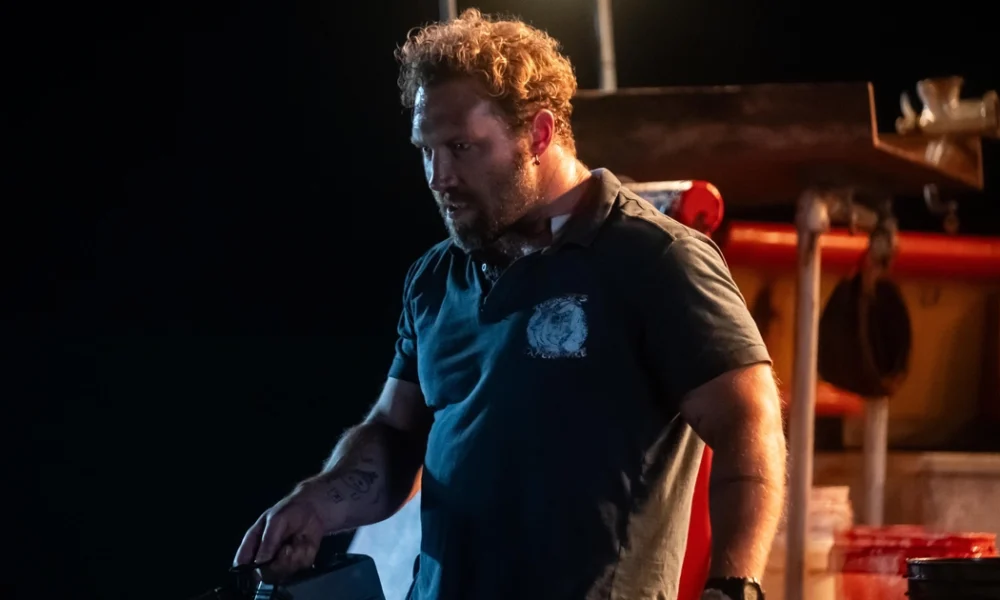 Jai Courtney em cena tensa à noite no filme Animais Perigosos (2025), usando camiseta escura em um barco pesqueiro iluminado por luz artificial.